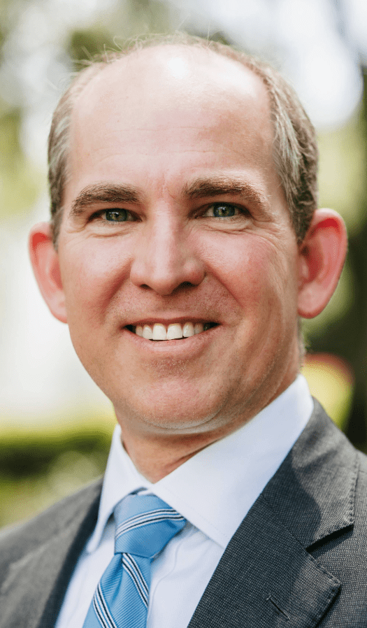 Smiling man in a suit with a blue striped tie stands outdoors against a blurred green background